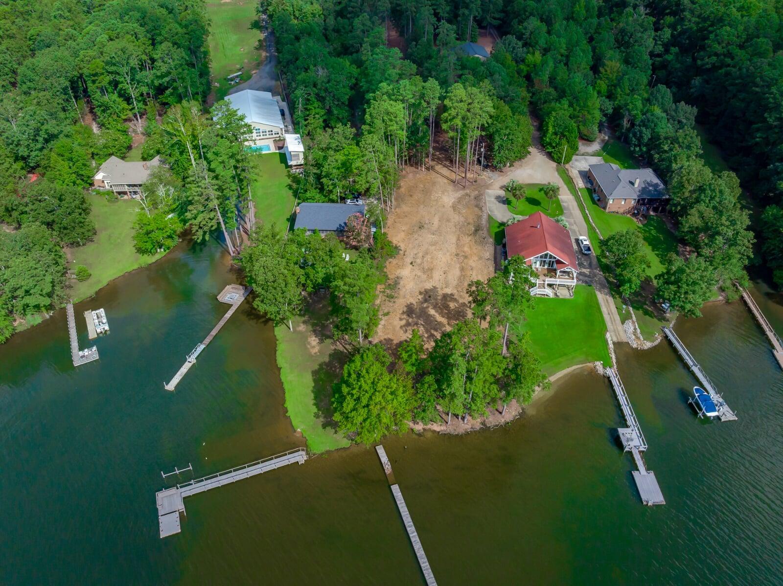 A cinematic aerial photograph showcasing a luxury waterfront property featuring three private boat docks, a large cleared lot, and a substantial home with a red roof beside a dark lake, emphasizing high-end real estate and development potential.